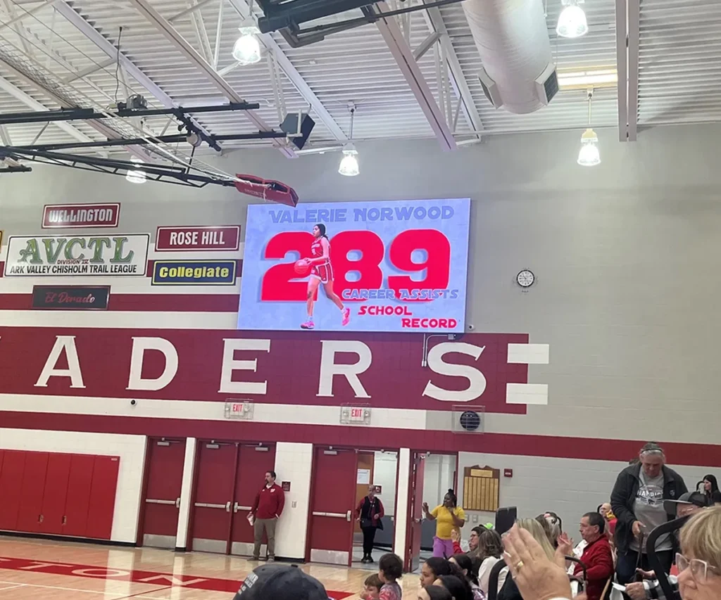 An LED electronic message center in the gym displays Valerie Norwood’s 2,289 career points school record alongside her photo. Spectators and students can be seen beneath the high-brightness display, some clapping and others watching the solid-state scoreboard screen.