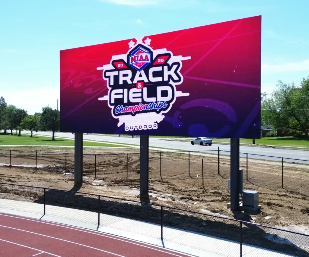 High-brightness LED electronic message center with aluminum housing near an athletic track displays "MIAA 2026 Track & Field Championships Outdoor" in large bold letters; a car drives by on the nearby road in the background.