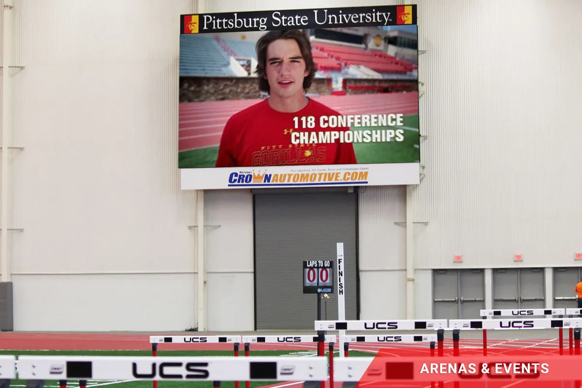 High-brightness LED electronic message center at an indoor track displays an image of a student with the text, "Pittsburg State University. 118 Conference Championships." Track hurdles and a commercial ARENAS & EVENTS aluminum sign appear in the foreground.