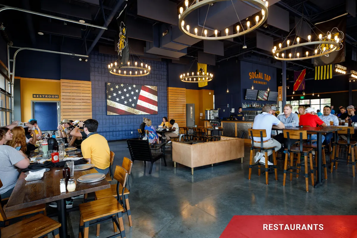 Modern restaurant interior with patrons dining; a high-brightness LED American flag display is mounted on the wall. Chandeliers hang overhead, and a bar area with solid-state electronic message center signage is visible in the background. The floor is polished concrete, creating an accessible, casual setting.