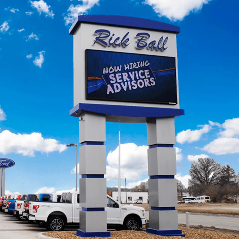 A tall LED electronic message center at Rick Ball car dealership displays "Now Hiring Service Advisors." Multiple white trucks are lined up in front beneath a bright blue sky with scattered clouds.