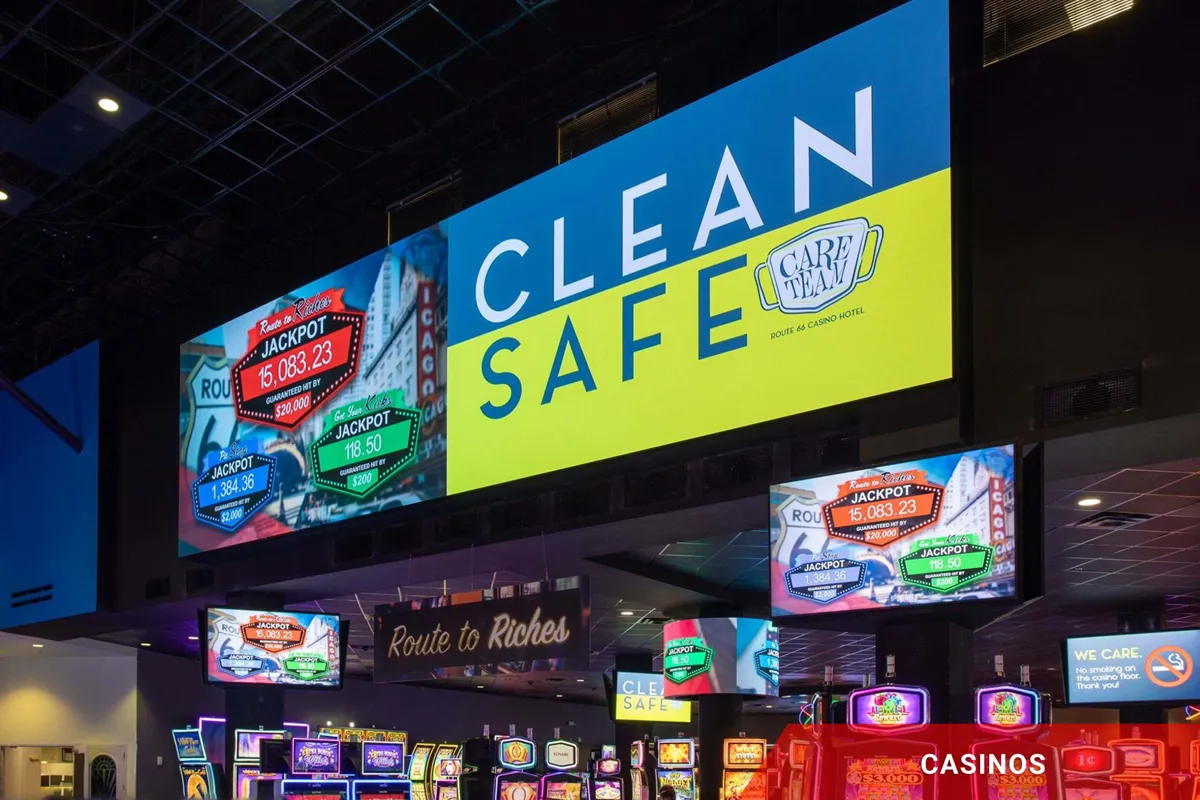 A casino interior featuring rows of slot machines and an overhead LED electronic message center displaying CLEAN SAFE in high-brightness, solid-state lettering, highlighting cleanliness and safety throughout the gaming area.
