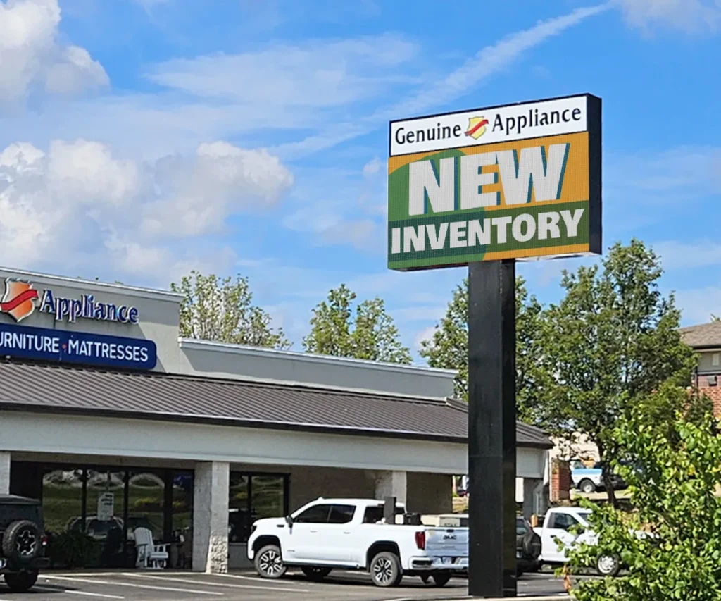 A retail store features an LED electronic message center displaying "Genuine Appliance NEW INVENTORY," situated before a parking lot filled with white vehicles and bordered by trees beneath a partly cloudy sky.