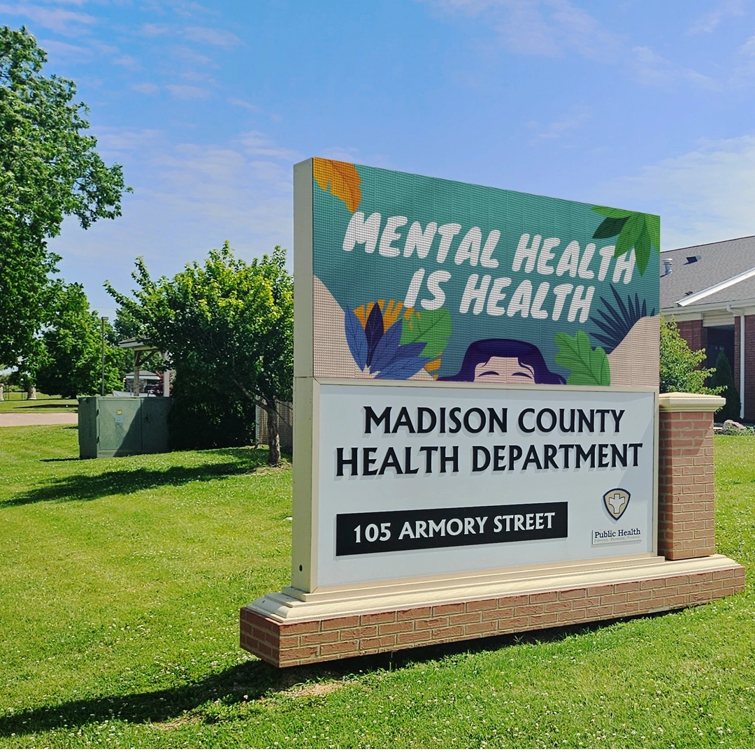STAX an outdoor LED sign with high-brightness display reads "Mental Health is Health," "Madison County Health Department," and "105 Armory Street." The electronic message center is positioned in front of a building, surrounded by greenery and set against a blue sky.