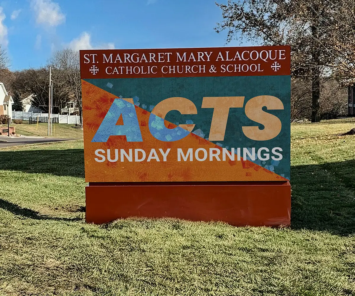 an LED electronic message center sign with a colorful display stands on grass, showing the text "ST. MARGARET MARY ALACOQUE CATHOLIC CHURCH & SCHOOL. ACTS SUNDAY MORNINGS" under a clear sky.