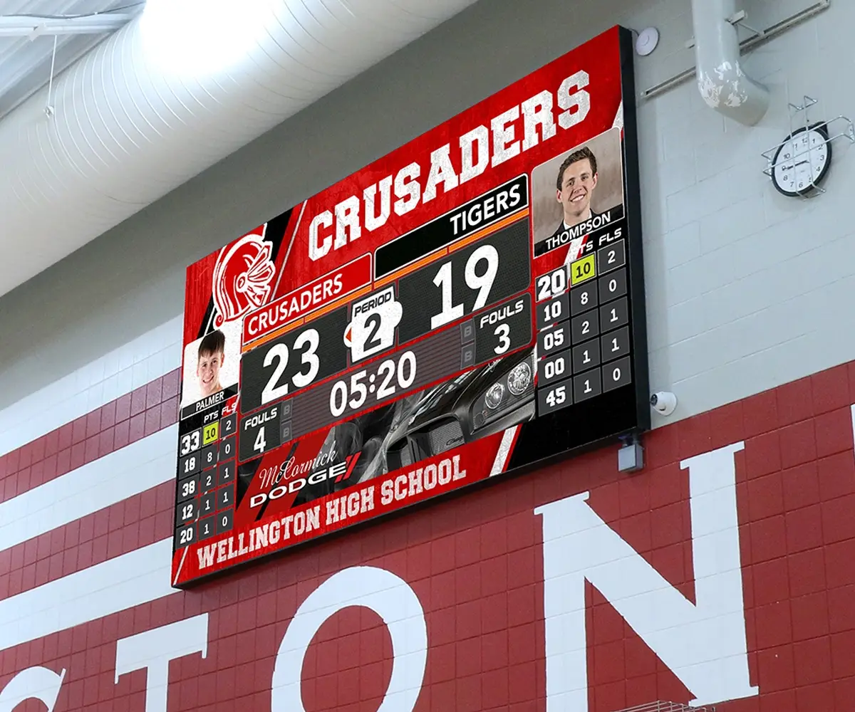 LED electronic message center in a gym displays Crusaders leading Tigers 23-19 during the second period with 5:20 remaining. Player stats and photos are visible on the screen. The scoreboard is installed above Wellington High School signage.
