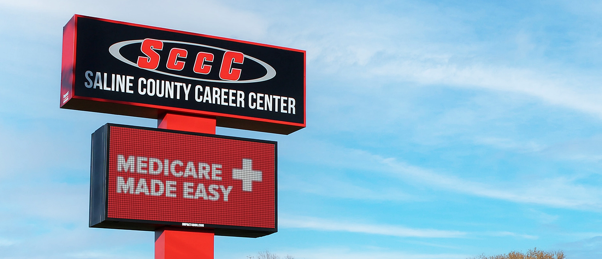 A red and black SCCC Saline County Career Center sign stands above an LED electronic message center displaying “Medicare Made Easy” alongside a white medical cross. The signage is set against a partly cloudy blue sky.