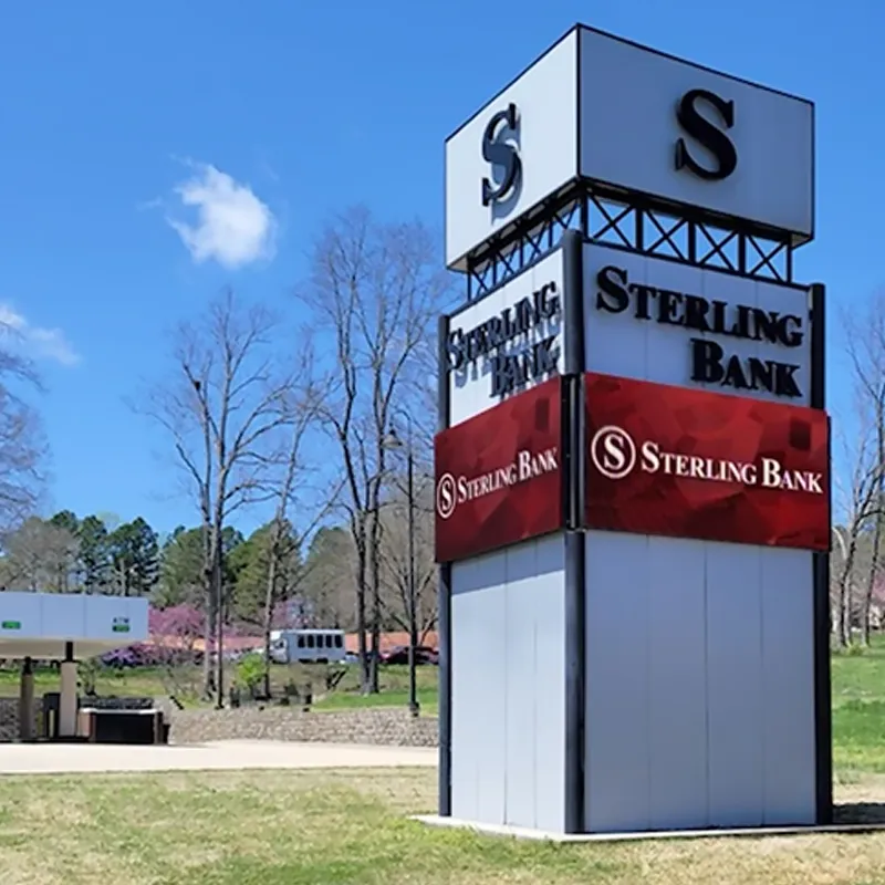 Sterling Bank’s tall commercial sign features high-brightness red and white panels, possibly LED or electronic message center, mounted on a grassy area next to a parking lot. Leafless trees and a partly cloudy blue sky are visible in the background.