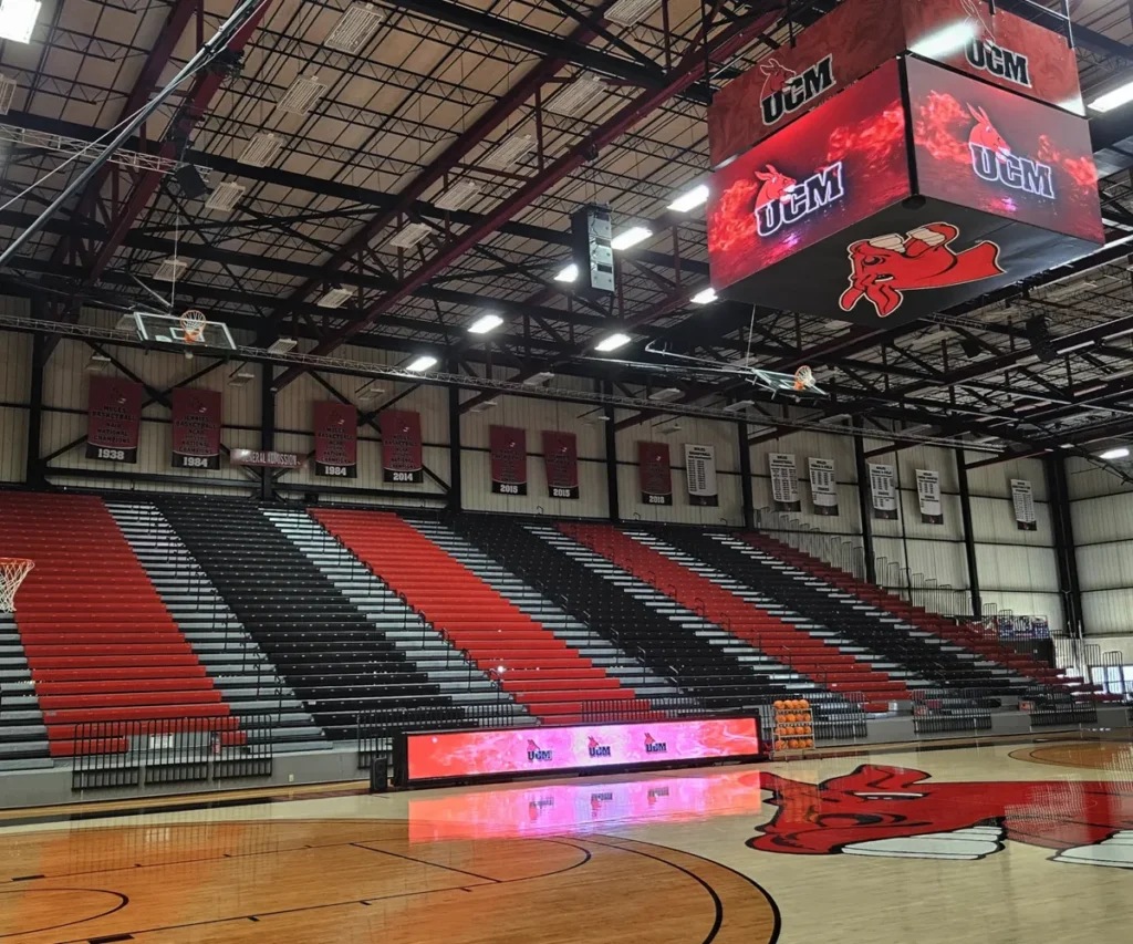 An empty gymnasium featuring red and black bleachers, a basketball court, wall-mounted banners, and a high-brightness solid-state LED electronic message center (EMC) scoreboard displaying UCM.