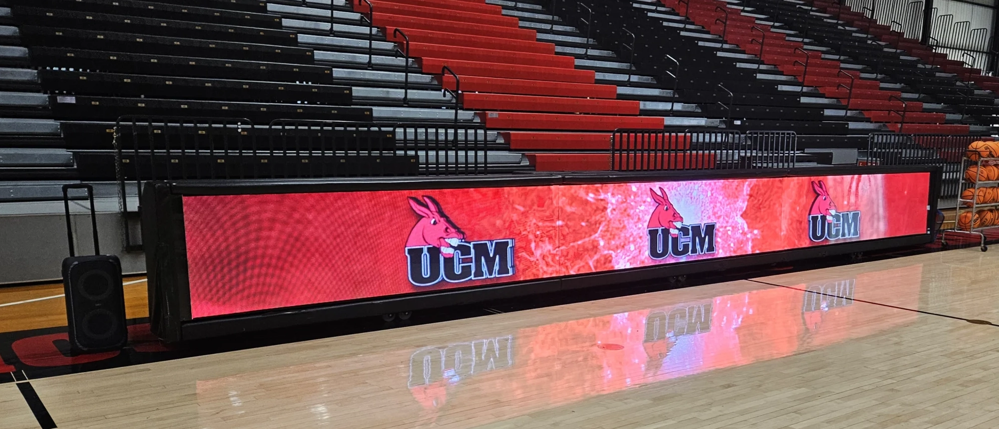 LED scorer’s table beside the University of Central Missouri basketball court showing repeated UCM lettering and red mule graphics in front of black and red bleachers.