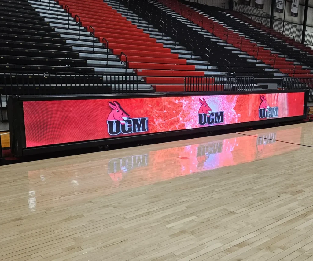A high-brightness solid-state LED electronic message center at a basketball court displays the UCM logo with a red mule head and text on a red background. Black and red aluminum bleachers are seen in the background.