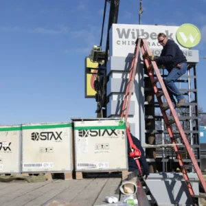 Worker on a ladder installing a sign at Wilbeck Chiropractic, with a crane and STAX shipping crates beside the sign structure.