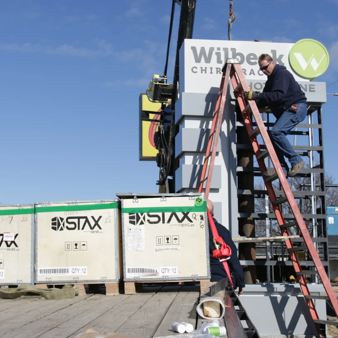 Worker on a ladder installing a sign at Wilbeck Chiropractic, with a crane and STAX shipping crates beside the sign structure.