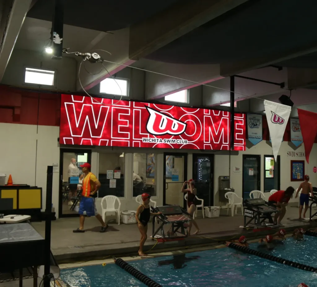 Swimmers prepare by an indoor pool beneath a high-brightness LED WELCOME sign for Wichita Swim Club. The electronic message center is mounted on the back wall, with people and chairs visible along the perimeter.
