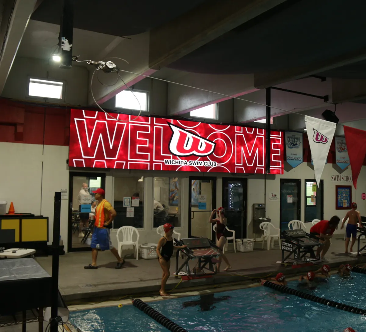 Swimmers prepare by an indoor pool beneath a high-brightness LED WELCOME sign for Wichita Swim Club. The electronic message center is mounted on the back wall, with people and chairs visible along the perimeter.