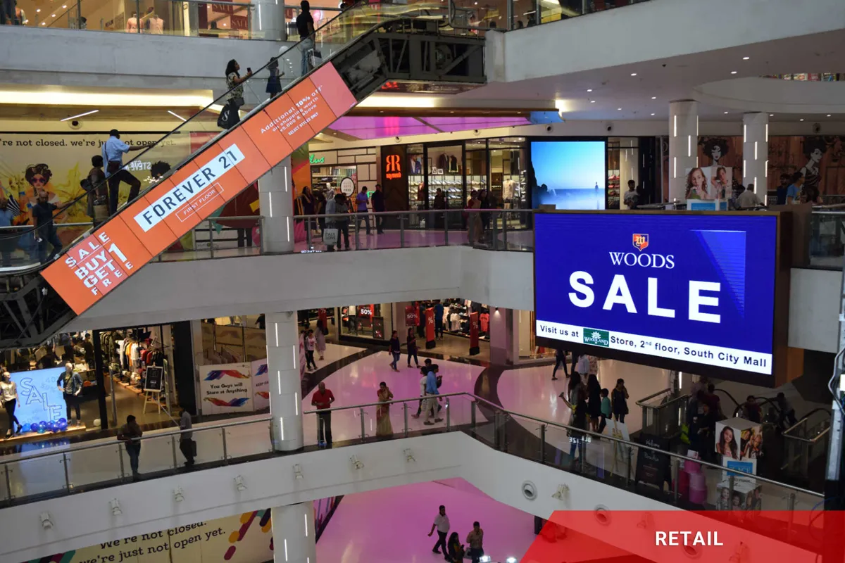 Shoppers navigate a multi-level mall featuring high-brightness LED signs for FOREVER 21 and WOODS. The scene includes people on escalators, electronic message centers above storefronts, and illuminated commercial signage throughout the vibrant retail environment.