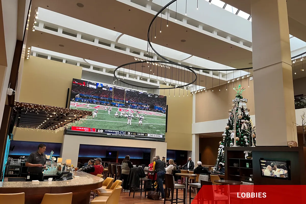 People gather in a spacious hotel lobby featuring a bar, large LED television displaying football, modern lighting fixtures, and a decorated Christmas tree. An electronic message center with the word "LOBBIES" appears in the corner, using high-brightness, solid-state display technology.