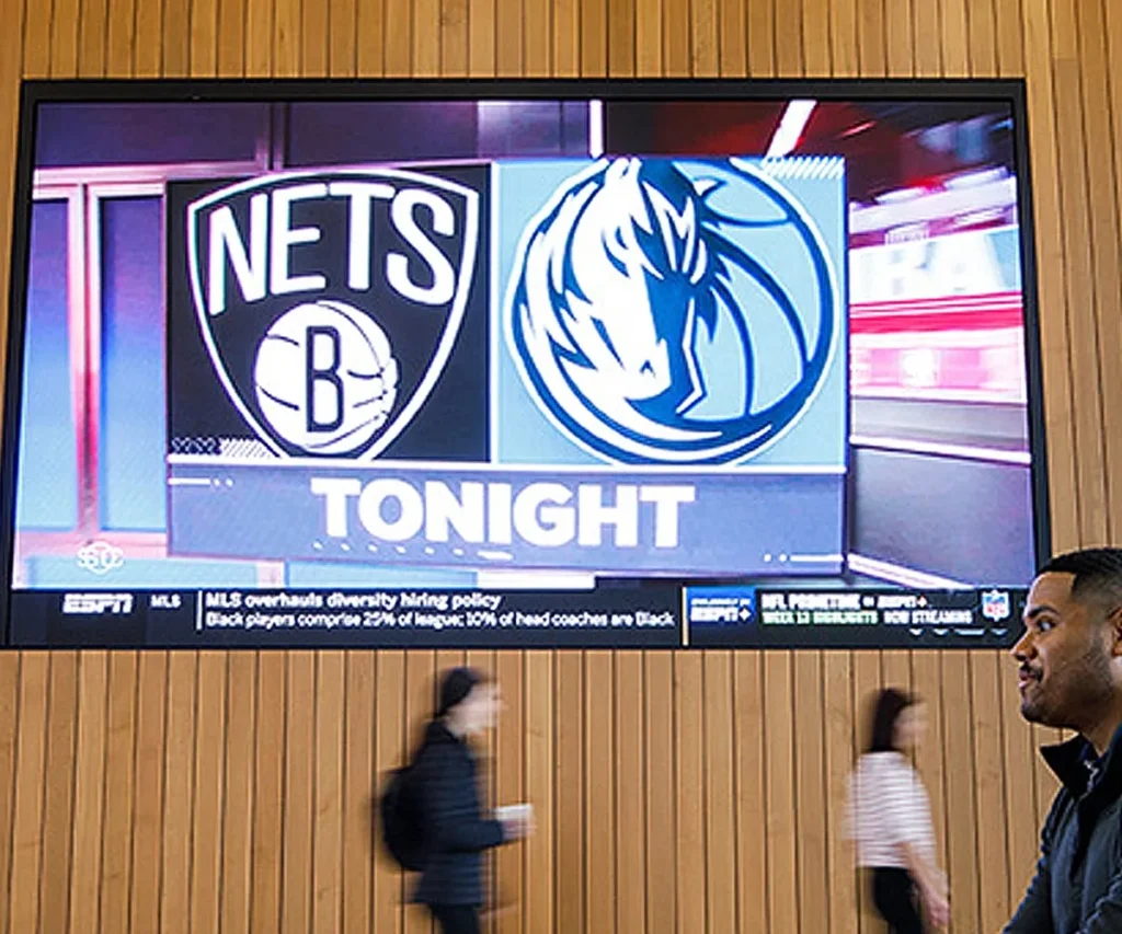 High-brightness LED electronic message center shows "Nets vs Mavericks Tonight" with team logos; pedestrians walk beneath the illuminated screen.