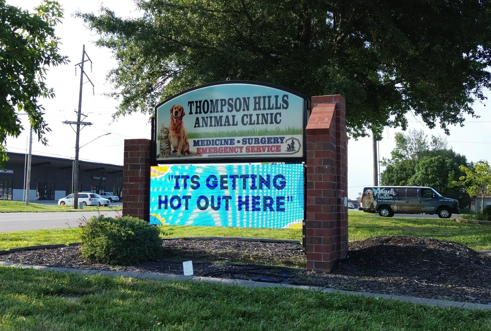 An LED electronic message center for Thompson Hills Animal Clinic displays “ITS GETTING HOT OUT HERE” in bright letters, with an aluminum frame. Background includes trees, a roadway, and several parked vehicles.