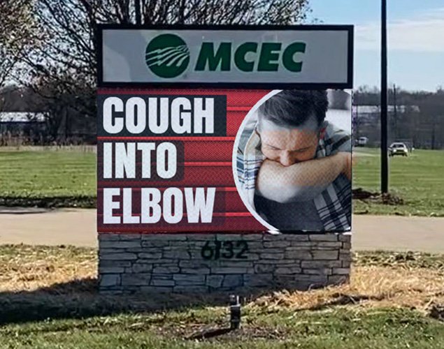 An outdoor LED sign with high-brightness display reads “COUGH INTO ELBOW” and features a graphic of a person coughing into their elbow. The solid-state electronic message center includes the MCEC logo at the top and is installed on a grassy area adjacent to a road.