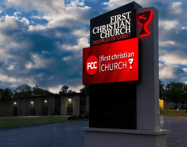 First Christian Church monument sign with white church lettering on a red LED display beneath a permanent church name and chalice topper at dusk.