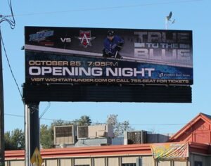 An LED electronic message center displays a hockey game promotion: October 25, 7:05 PM Opening Night with team logos, a hockey player image, and ticket information. Venue name and contact number appear beneath the event details on the signage.