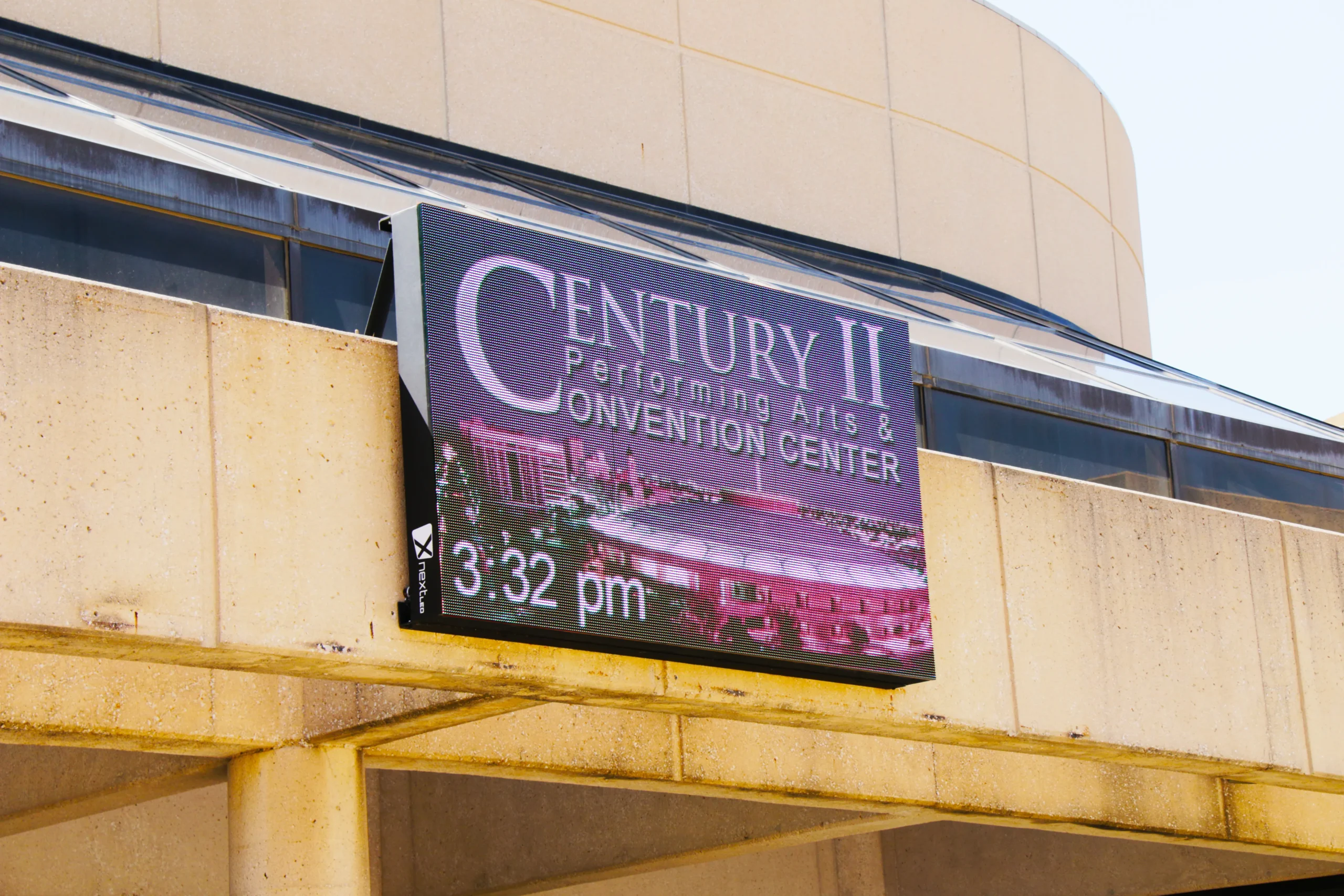 Wall-mounted LED display on the Century II Convention Center showing its name, a purple graphic, and 3:32 pm beneath the curved building facade.