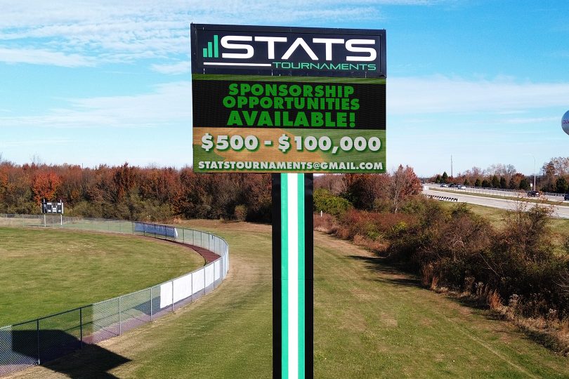 High-brightness LED sign in park promotes STATS Tournaments, sponsorships $500–$100k; contact info appears. Trees, water tower behind.