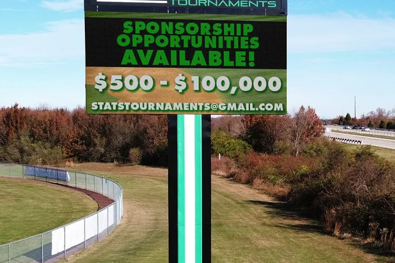 A tall LED electronic message center sign displays: STATS TOURNAMENTS, Sponsorship opportunities available $500–$100,000, and statstournaments@gmail.com. The solid-state sign is installed on an aluminum pole near a grassy field and a road lined with trees.