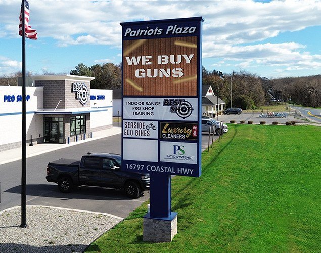 Patriots Plaza roadside monument shows We Buy Guns on its LED display above tenant panels beside the road and businesses.