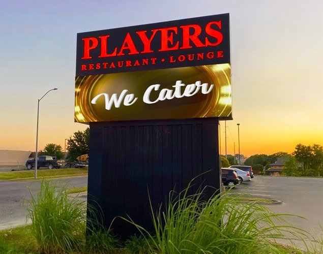 Players Restaurant and Lounge monument shows We Cater on its illuminated LED display at sunset, with plants around the sign's base.