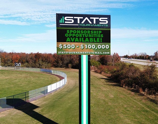 STATS Sports Complex pole sign shows sponsorship opportunities on its LED display beside landscaped athletic grounds and a curved path.
