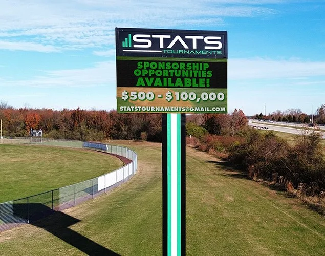 STATS Sports Complex pole sign shows sponsorship opportunities on its LED display beside landscaped athletic grounds and a curved path.